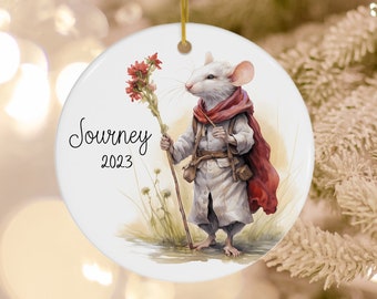May include: White ceramic Christmas ornament featuring a white mouse wearing a red and white cloak, holding a stick with a red flower on top. The ornament has the text "Journey 2023" on it.