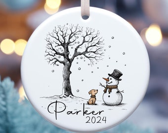 May include: White ceramic Christmas ornament with a black and white illustration of a dog and snowman in a snowy landscape with a tree. The ornament is personalized with the name "Parker" and the year "2024".