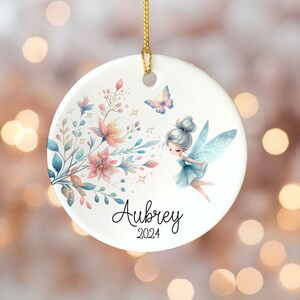 May include: A white ceramic ornament with a blue fairy with wings, flowers, and butterflies. The ornament is personalized with the name "Aubrey" and the year "2024".