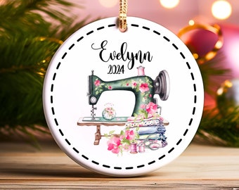May include: A white ceramic ornament with a green sewing machine with pink flowers, a spool of thread, and fabric. The ornament has a white border with a black dashed line and the text "Evelynn 2024".