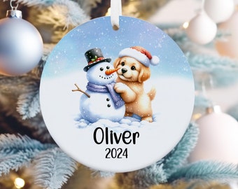 May include: A round, white Christmas ornament with a blue background. A cartoon puppy wearing a Santa hat is hugging a snowman. The ornament is personalized with the name "Oliver" and the year "2024".