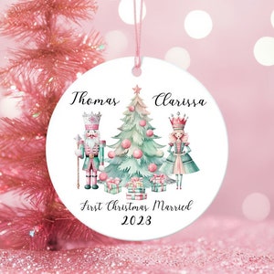 May include: A white round Christmas ornament with a watercolor illustration of a nutcracker and a sugar plum fairy standing on either side of a Christmas tree. The ornament is personalized with the names "Thomas" and "Clarissa" and the text "First Christmas Married 2023".