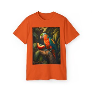 May include: Orange t-shirt featuring a colourful parrot eating fruit on a branch in a jungle setting. The text "Guatemala" is printed on the shirt.