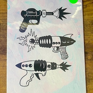 May include: Three black and white illustrations of toy ray guns on a white background with a light blue and pink marbled pattern. The ray guns have different designs and are all pointing to the right.