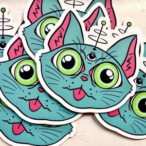 Könnte beinhalten: Ein Stapel blauer und rosa Katzenaufkleber mit Kulleraugen und einem dritten Auge auf der Stirn. Jede Katze hat eine rosa Zunge herausgestreckt und eine spiralförmige Antenne auf dem Kopf.
