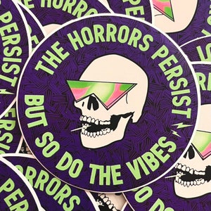 May include: A round sticker with a skull wearing green and pink sunglasses. The text on the sticker reads "The Horrors Persist But So Do The Vibes".