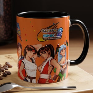 May include: A ceramic coffee mug with a black handle and a bright orange background. The mug features a colorful illustration of characters from the video game Capcom vs. SNK 2: Mark of the Millennium 2001. The text "CAPCOM VS. SNK 2 MARK OF THE MILLENNIUM 2001" is printed on the mug.