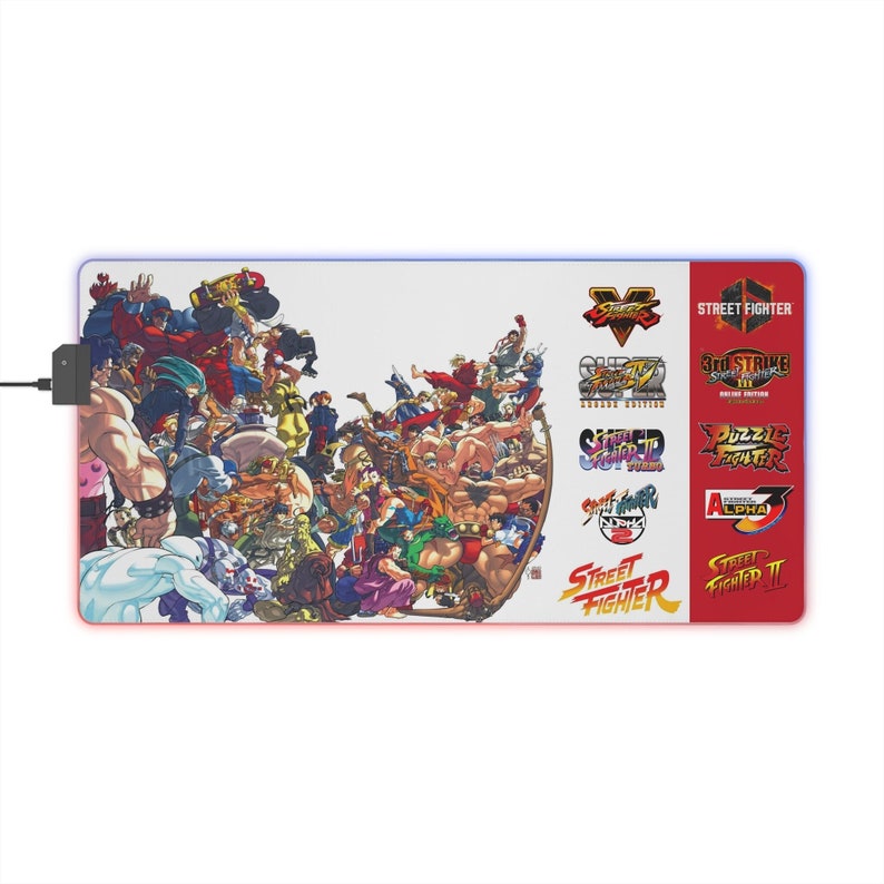 May include: Rectangular gaming mouse pad featuring a vibrant illustration of Street Fighter characters. The pad has a white background with a red border and includes various Street Fighter logos. A black USB connector is attached.