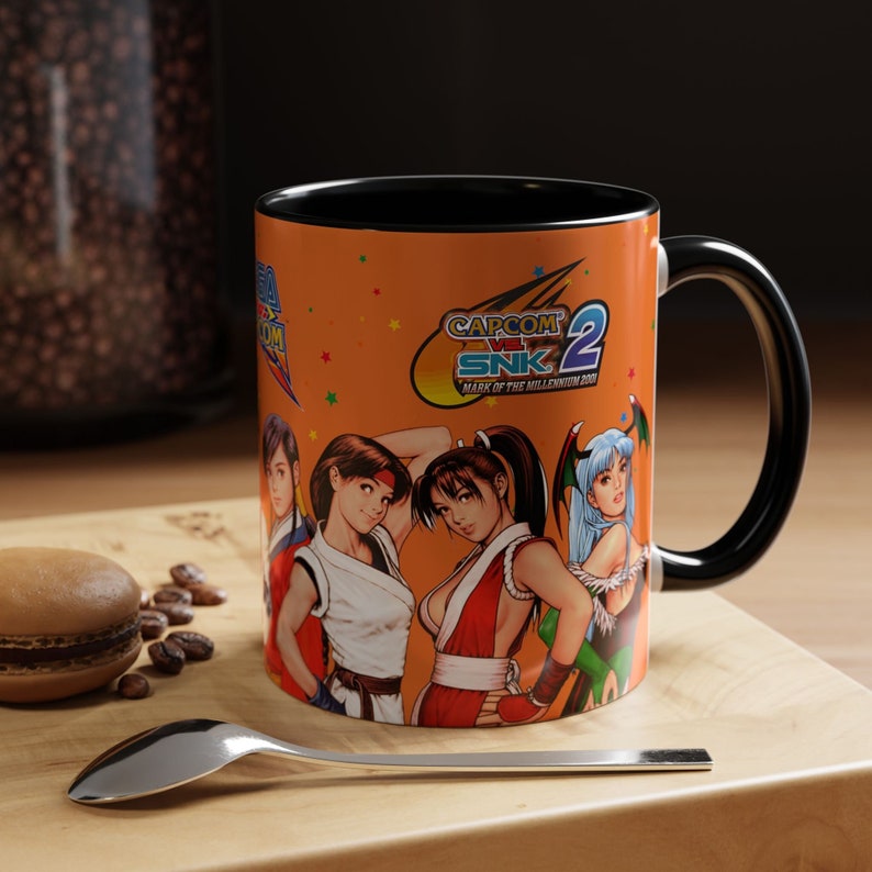 May include: A ceramic coffee mug with a black handle and a bright orange background. The mug features a colorful illustration of characters from the video game Capcom vs. SNK 2: Mark of the Millennium 2001. The text "CAPCOM VS. SNK 2 MARK OF THE MILLENNIUM 2001" is printed on the mug.