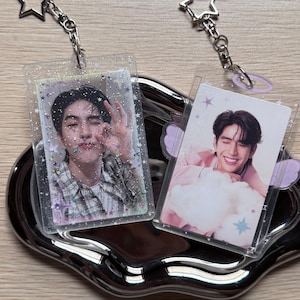 Pode incluir: Dois porta-retratos retangulares de acrílico transparente com chaveiros de metal em forma de estrela. O suporte esquerdo contém a foto de uma pessoa fazendo um gesto de ok com glitter. O direito mostra uma pessoa sorrindo com asas de cor lilás.
