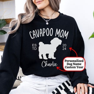 May include: Black sweatshirt with white text that reads "CAVAPOO MOM" and a white silhouette of a dog. The text "EST TD" and "20 24" are also printed on the sweatshirt. The text "Charlie" is printed below the dog silhouette. The text "Personalized Dog Name Custom Year" is printed on a white rectangle.