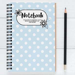 May include: A spiral-bound notebook with a light blue polka dot cover and the word "Notebook" in a decorative font. The notebook is accompanied by a black pencil. The cover also features a floral design and a Bible verse.