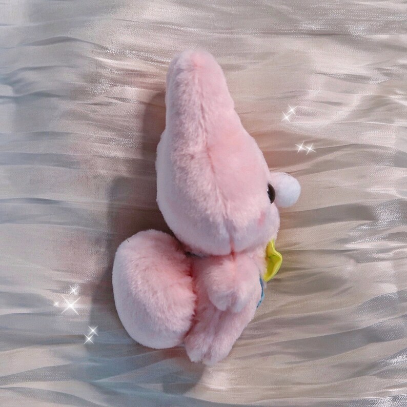 May include: Pink plush toy shaped like a cartoon character with a yellow bow. The toy is sitting on a white textured surface.
