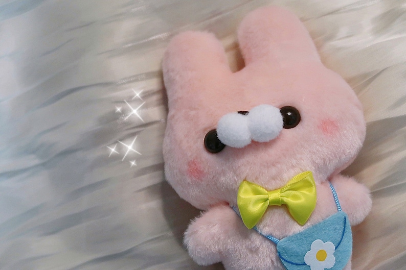May include: A pink plush bunny with a yellow bow and a blue purse with a white flower. The bunny has large white eyes and black pupils.