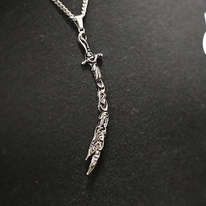 May include: A silver necklace with a detailed sword pendant. The pendant is intricately designed with Arabic calligraphy.