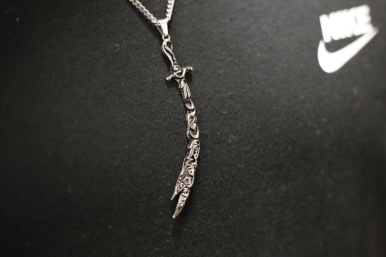 May include: A silver necklace with a detailed sword pendant. The pendant is intricately designed with Arabic calligraphy.