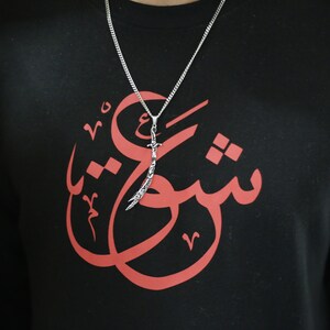 May include: A black sweatshirt with a red Arabic calligraphy design and a silver sword pendant necklace.
