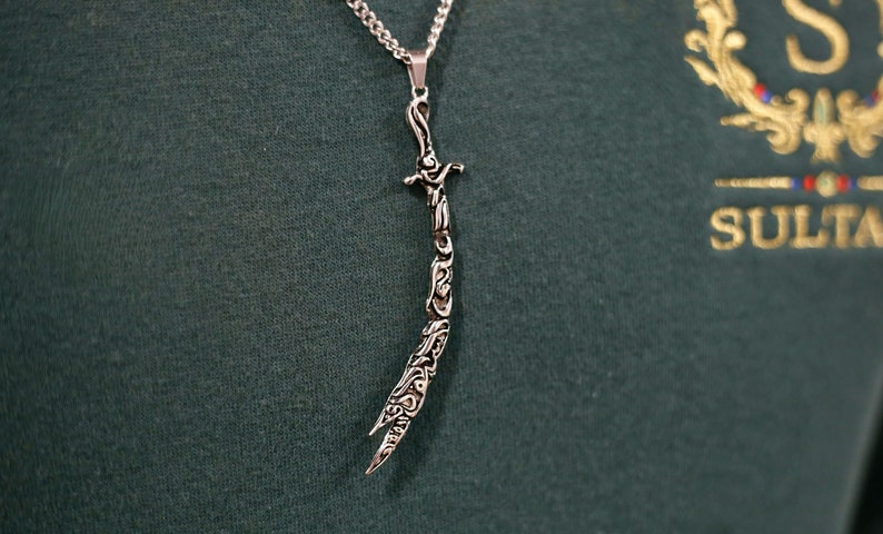May include: A silver necklace with a pendant shaped like a sword. The sword is intricately designed with Arabic calligraphy.