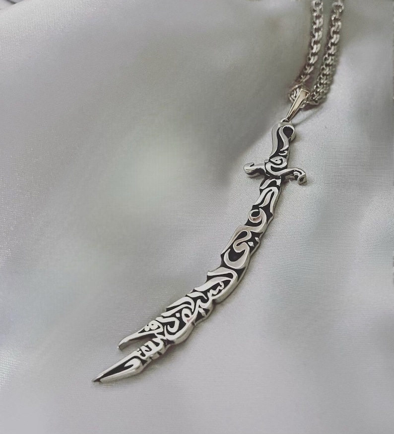 May include: A silver necklace with a sword pendant. The sword is intricately designed with Arabic calligraphy.