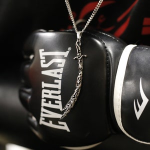 May include: A silver chain necklace with a detailed sword pendant. The necklace is worn over a black Everlast boxing glove.
