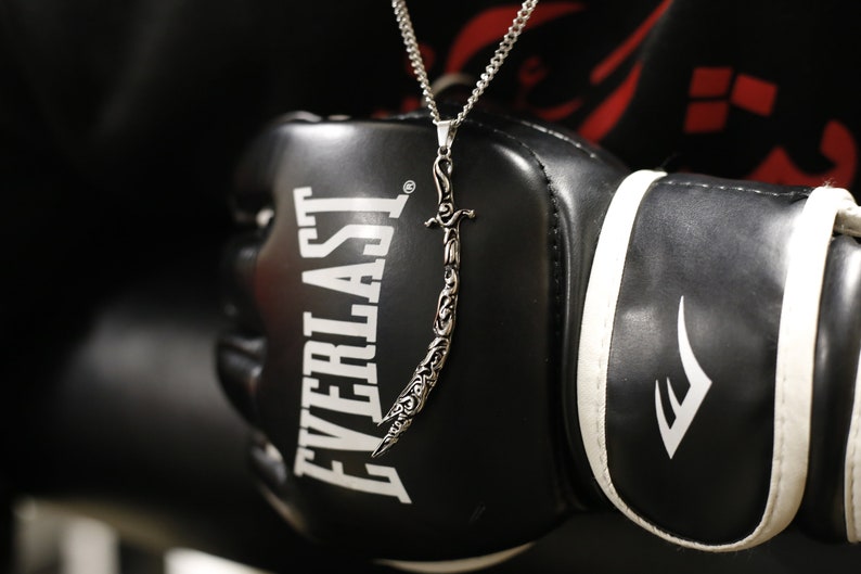 May include: A silver chain necklace with a detailed sword pendant. The necklace is worn over a black Everlast boxing glove.