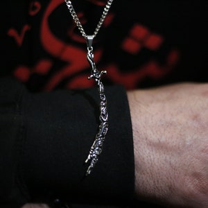 May include: A silver chain necklace with a pendant shaped like a sword. The sword is detailed with intricate carvings.