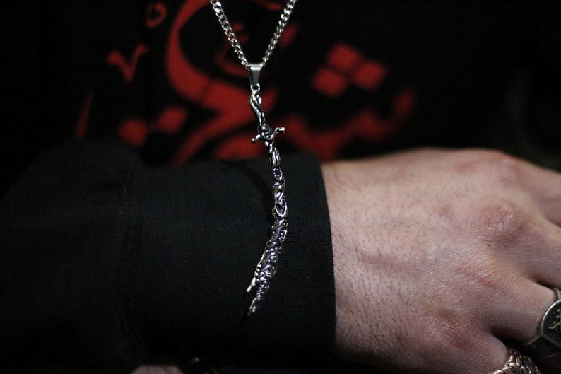 May include: A silver chain necklace with a pendant shaped like a sword. The sword is detailed with intricate carvings.