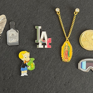 May include: A collection of ten enamel pins featuring various designs, including a feather with an American flag, a bottle of tequila, a marijuana leaf, a ski goggle, a Mexican flag, a religious icon, a cartoon character holding a bag of money, and the letters "LA".