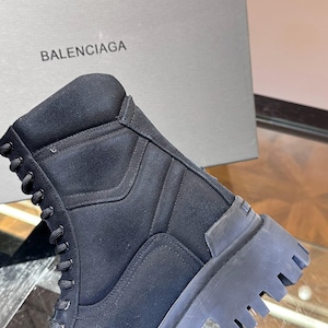 May include: Black combat boot with a thick, ridged sole. The boot features a lace-up design and a padded collar. The word "BALENCIAGA" is printed on the box in the background.