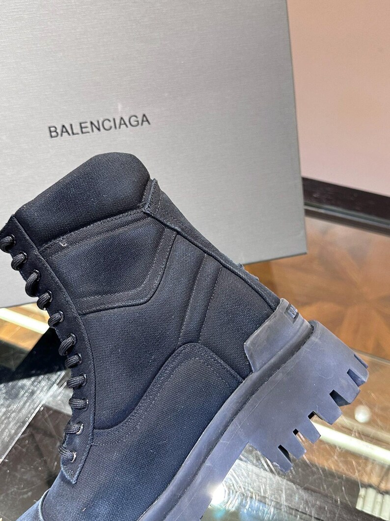May include: Black combat boot with a thick, ridged sole. The boot features a lace-up design and a padded collar. The word "BALENCIAGA" is printed on the box in the background.