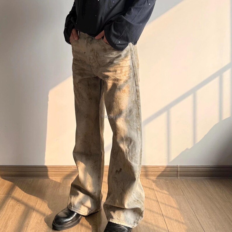 May include: Full-length denim jeans featuring a light tan and beige wash with a distressed, mottled appearance. The trousers have a straight-leg cut, a standard waistband with belt loops, and visible stitching details throughout the fabric surface.