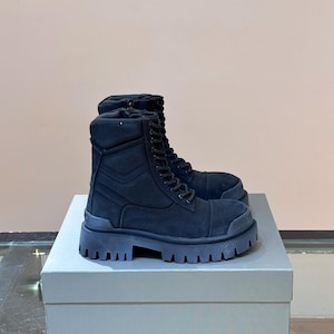 May include: A pair of black combat boots with thick, lug soles. The boots have a lace-up design and are made of a suede-like material. They are displayed on a gray box.