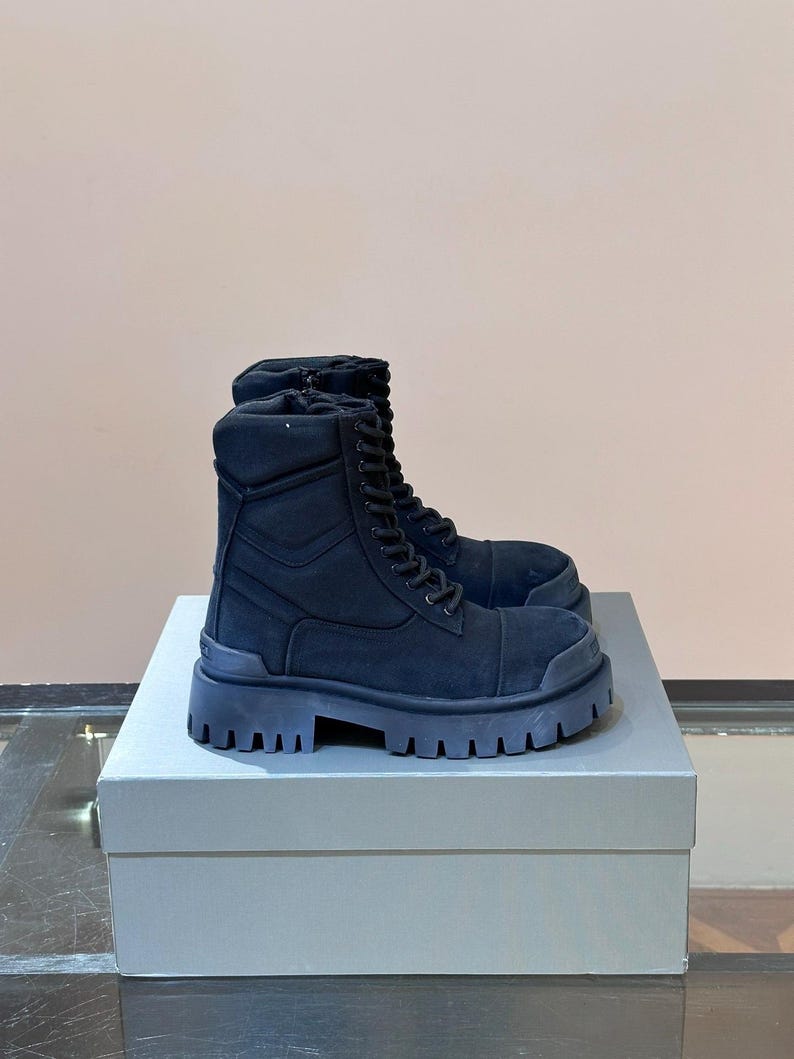 May include: A pair of black combat boots with thick, lug soles. The boots have a lace-up design and are made of a suede-like material. They are displayed on a gray box.