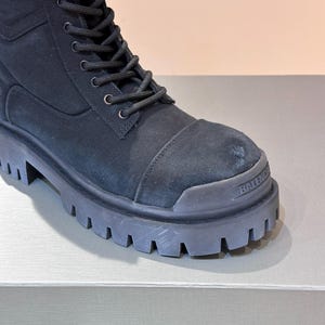 May include: Black combat boot with a thick, lugged sole. The boot features a canvas upper with a lace-up closure and a reinforced toe cap. The brand name "BALENCI" is visible on the toe.
