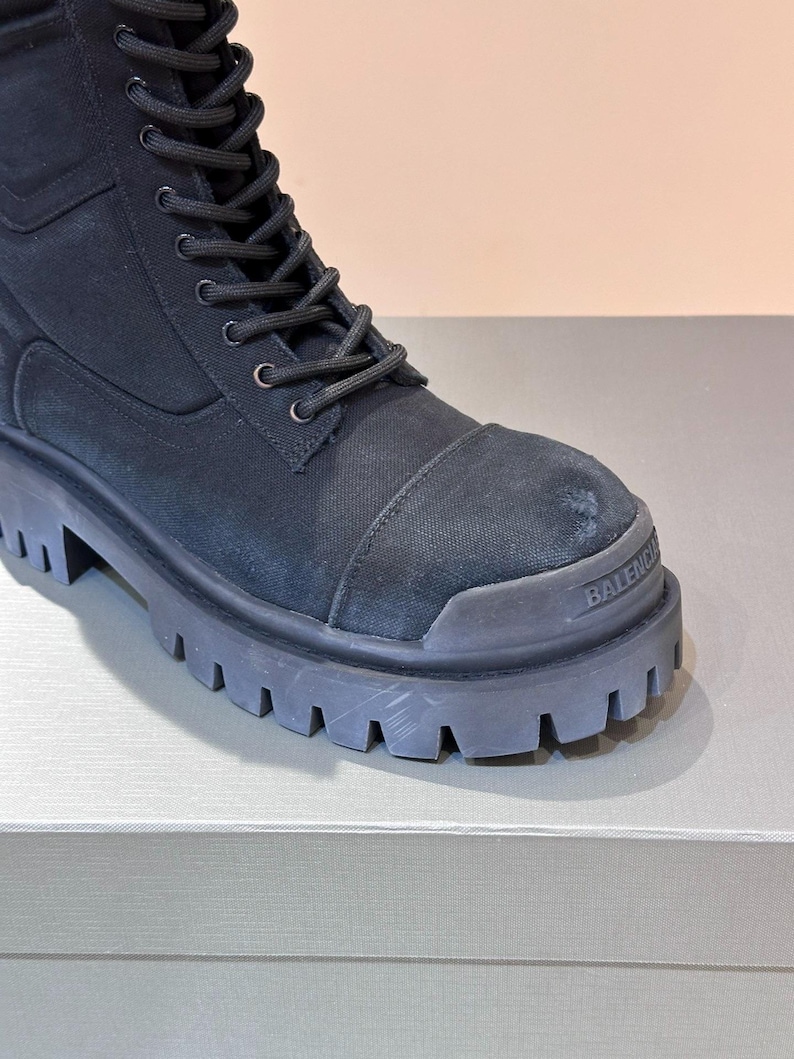 May include: Black combat boot with a thick, lugged sole. The boot features a canvas upper with a lace-up closure and a reinforced toe cap. The brand name "BALENCI" is visible on the toe.