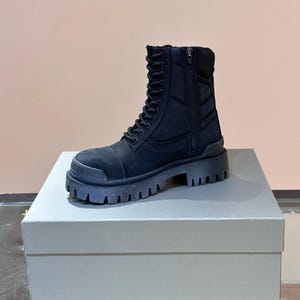May include: A black combat boot with a thick, lugged sole. The boot features a lace-up front, a side zipper, and a padded collar. The boot is made of a matte material and sits on a platform. The boot is displayed on a white box.