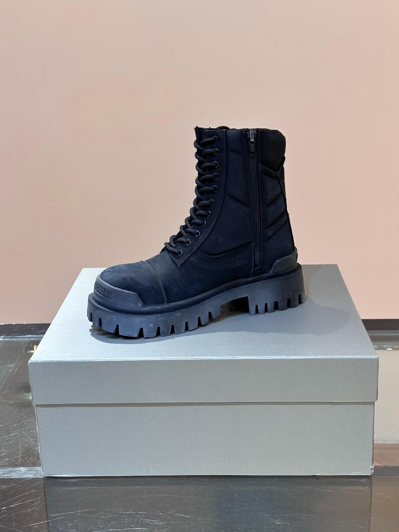 May include: A black combat boot with a thick, lugged sole. The boot features a lace-up front, a side zipper, and a padded collar. The boot is made of a matte material and sits on a platform. The boot is displayed on a white box.