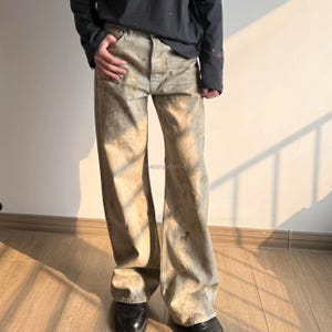May include: Wide-leg denim jeans featuring a distressed, mottled beige and grey wash with visible brown staining throughout. The trousers have a standard button fly closure, five-pocket construction, and a loose, relaxed fit. The text happywhale is visible.