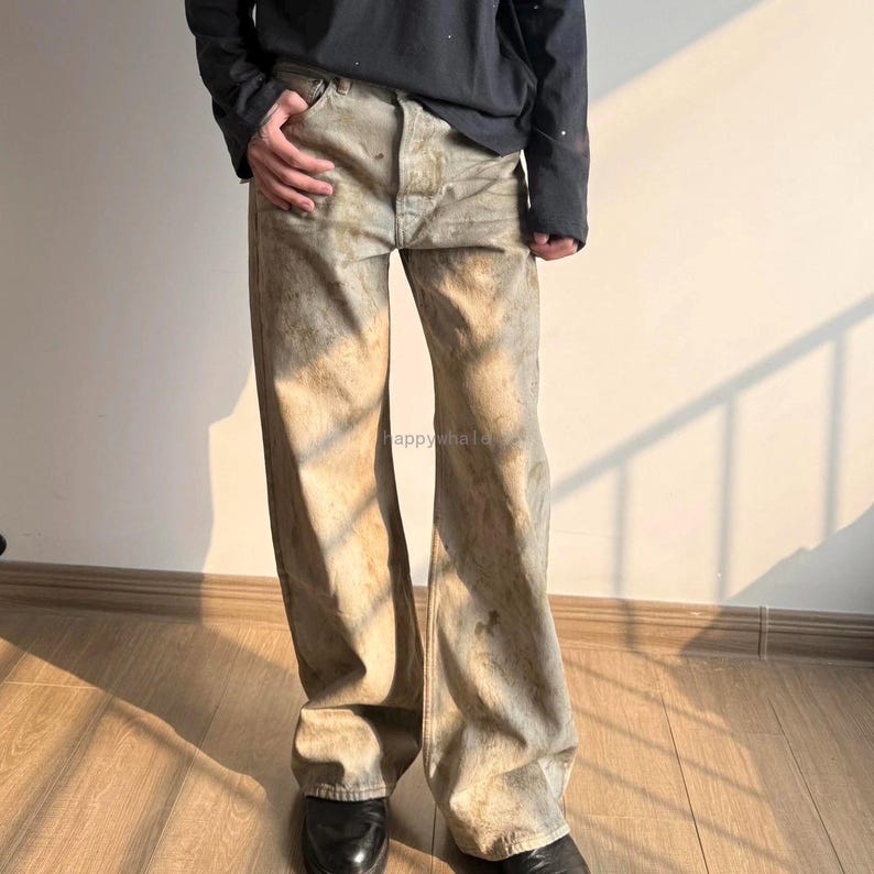 May include: Wide-leg denim jeans featuring a distressed, mottled beige and grey wash with visible brown staining throughout. The trousers have a standard button fly closure, five-pocket construction, and a loose, relaxed fit. The text happywhale is visible.