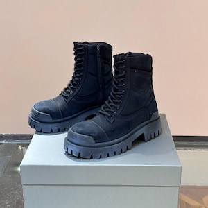 May include: A pair of black combat boots with thick, lug soles. The boots have a lace-up design with a side zipper and are made of a suede-like material. They are displayed on a light gray box.