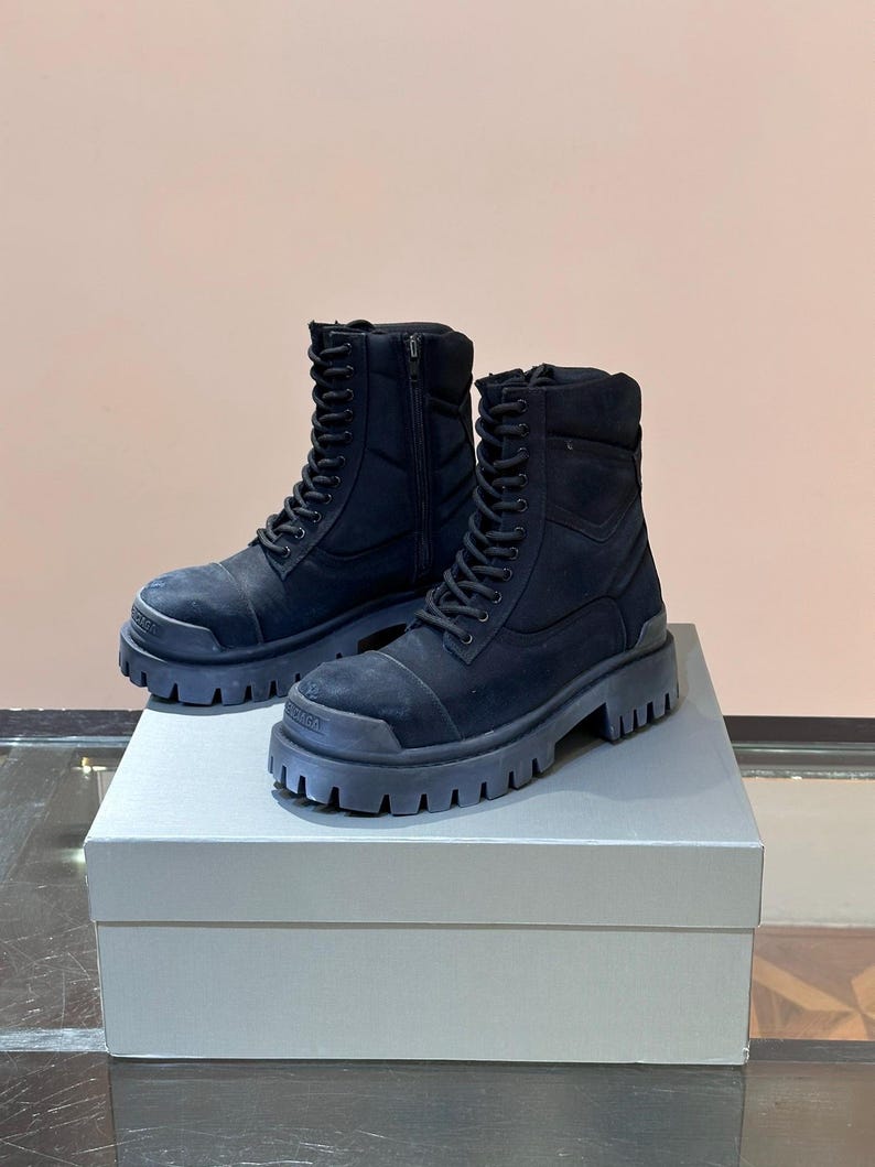 May include: A pair of black combat boots with thick, lug soles. The boots have a lace-up design with a side zipper and are made of a suede-like material. They are displayed on a light gray box.