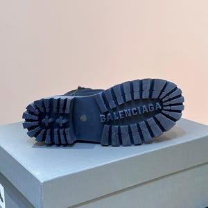 May include: A black boot with a thick, ridged sole. The word "BALENCIAGA" is embossed on the sole. The boot is resting on a gray box.