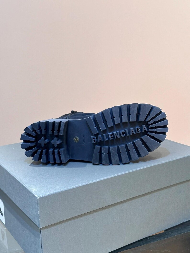 May include: A black boot with a thick, ridged sole. The word "BALENCIAGA" is embossed on the sole. The boot is resting on a gray box.