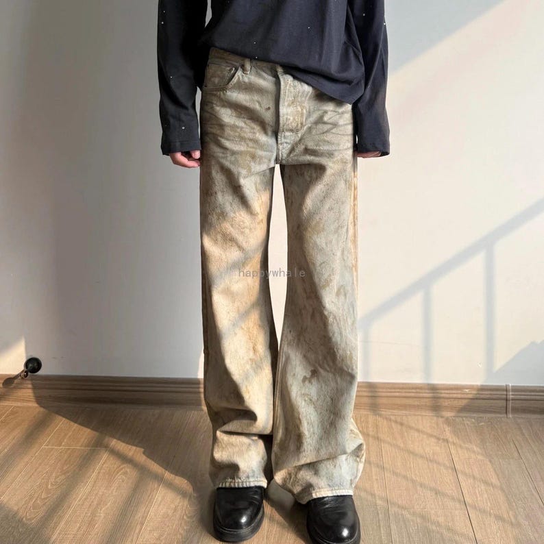 May include: Wide-leg denim jeans featuring a distressed, mottled beige and light blue wash with a button fly closure. The fabric displays a weathered, uneven texture throughout the legs, paired with a plain black long-sleeved shirt.
