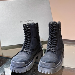 May include: Black combat boots with thick soles and lace-up closures. The boots have a rugged design and the brand name "BALENCIAGA" is visible on the soles. A gray box with the brand name is in the background.