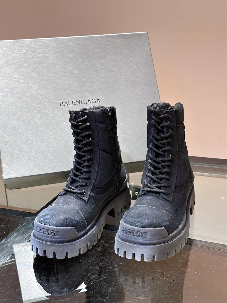 May include: Black combat boots with thick soles and lace-up closures. The boots have a rugged design and the brand name "BALENCIAGA" is visible on the soles. A gray box with the brand name is in the background.