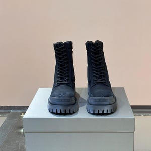 May include: A pair of black combat boots with thick, gray soles. The boots have laces and zippers, and the brand name "BALENCIAGA" is visible on the front of the soles. The boots are displayed on a white box.