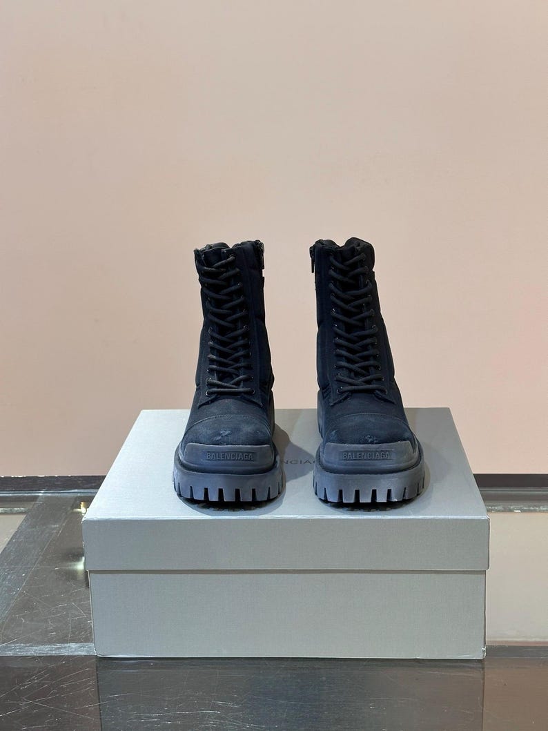 May include: A pair of black combat boots with thick, gray soles. The boots have laces and zippers, and the brand name "BALENCIAGA" is visible on the front of the soles. The boots are displayed on a white box.