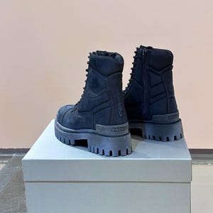 May include: A pair of black combat boots with thick, lug soles. The boots have a lace-up design and a side zipper. The brand name "BALENCIAGA" is visible on the heel. The boots are displayed on a light gray box.