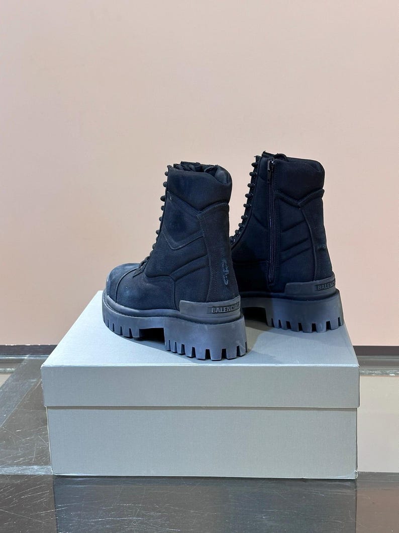 May include: A pair of black combat boots with thick, lug soles. The boots have a lace-up design and a side zipper. The brand name "BALENCIAGA" is visible on the heel. The boots are displayed on a light gray box.